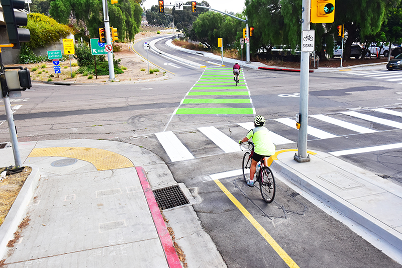 Two people riding bikes use new separated bike lanes and bike crossing paths as they travel on 19th Street. A freeway onramp, stop lights, and a car waiting to cross the intersection are visible.