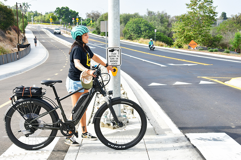 A woman wearing a helmet and walking her bike pushes the crosswalk button to cross Pershing Drive. A car, a jogger, and a motorcycle rider are visible in the background.