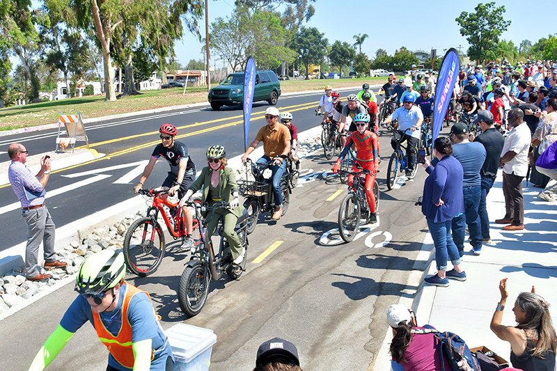 SANDAG Vice Chair and San Diego City Council President Sean Elo-Rivera and Executive Director of the San Diego County Bicycle Coalition Chloé Lauer lead the way as dozens of bike riders follow behind on the newly opened Pershing Bikeway and many others look on and cheer.