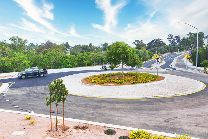 A black truck exits the new roundabout at Redwood Street and Pershing Drive. The center of the large roundabout is planted with trees and ground cover and several pedestrian-crossing signs, sidewalks, and bike lanes are visible in the distance.