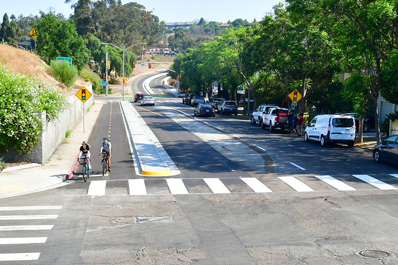 A view of two people riding bikes on a separated bikeway on 19th Street at C Street in San Diego. Parked cars, crosswalks, and freeway onramps are visible around them.