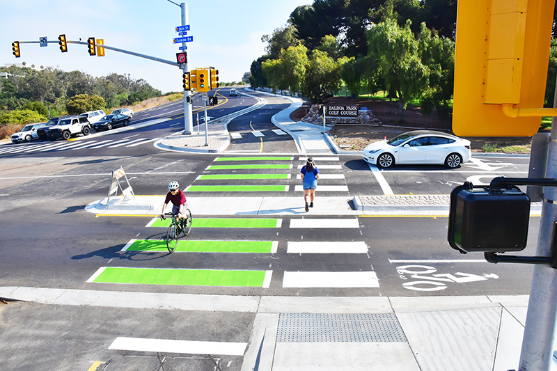 A southward view of Pershing Drive shows a person riding a bike  northbound in a separated bikeway and a person walking the other way in a pedestrian lane. Balboa Park Golf Course is in the background at the intersection of 26th Street and Florida Drive.