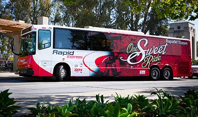 A red and white Rapid Express bus parked at a bus stop with surrounding tall green trees and a small cement archway in the background. The back half profile of the bus is covered in a red brick graphic and the words "One Sweet Ride" text on top.