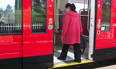 A middle-aged women is shown boarding a trolley with yellow stripping of tactile paving on the edge of the sidewalk that meets the trolley.